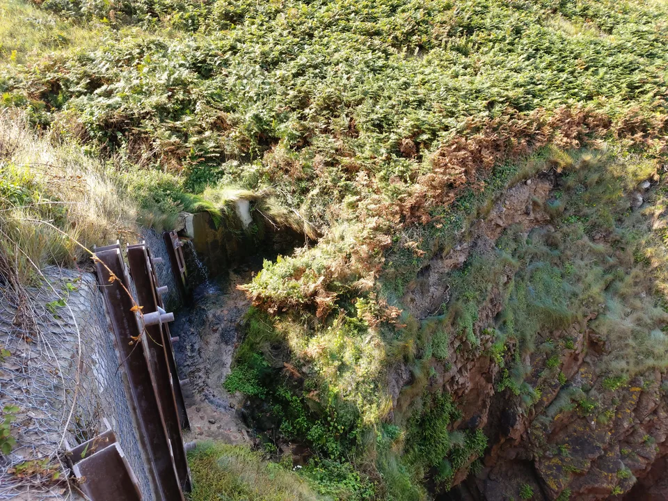 Water runs down a steep vegetated cliff beside metal structures at Plémont's coastal beach access.