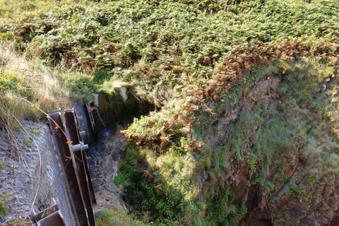 Water runs down a steep vegetated cliff beside metal structures at Plémont's coastal beach access.