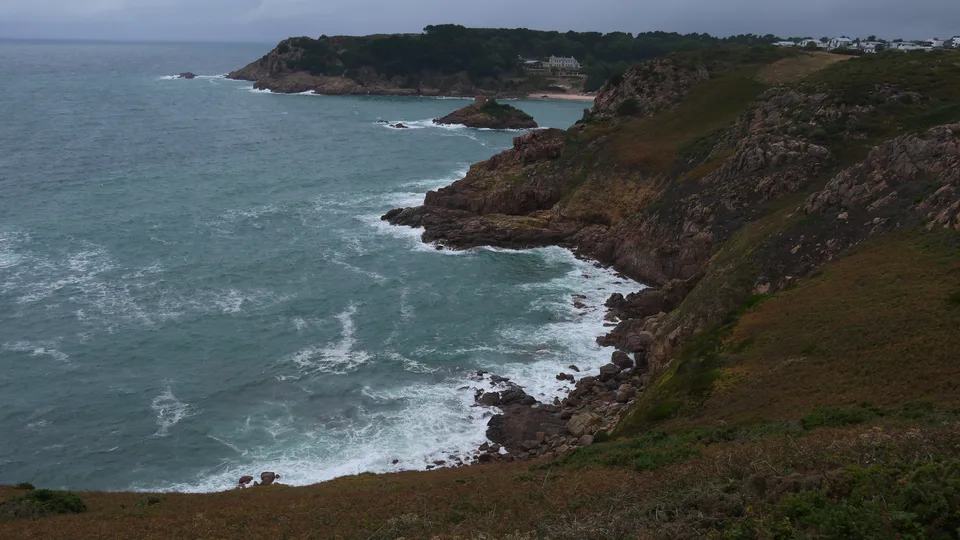 White waves break against a rugged rocky coastline topped with green trees beneath a cloudy sky.