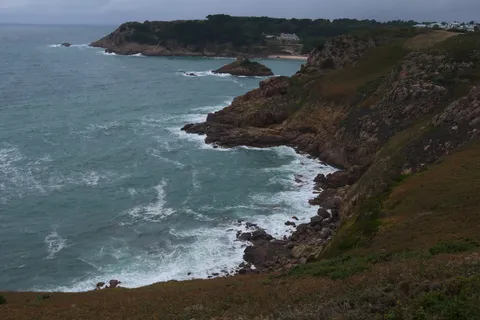 White waves break against a rugged rocky coastline topped with green trees beneath a cloudy sky.