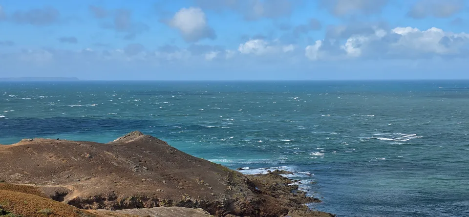 Small white waves break beside a dark rocky headland at Plémont beneath a partly cloudy blue sky.