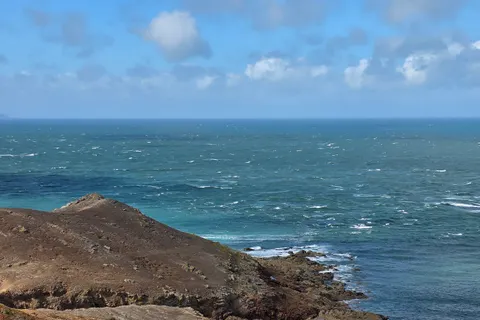 Small white waves break beside a dark rocky headland at Plémont beneath a partly cloudy blue sky.