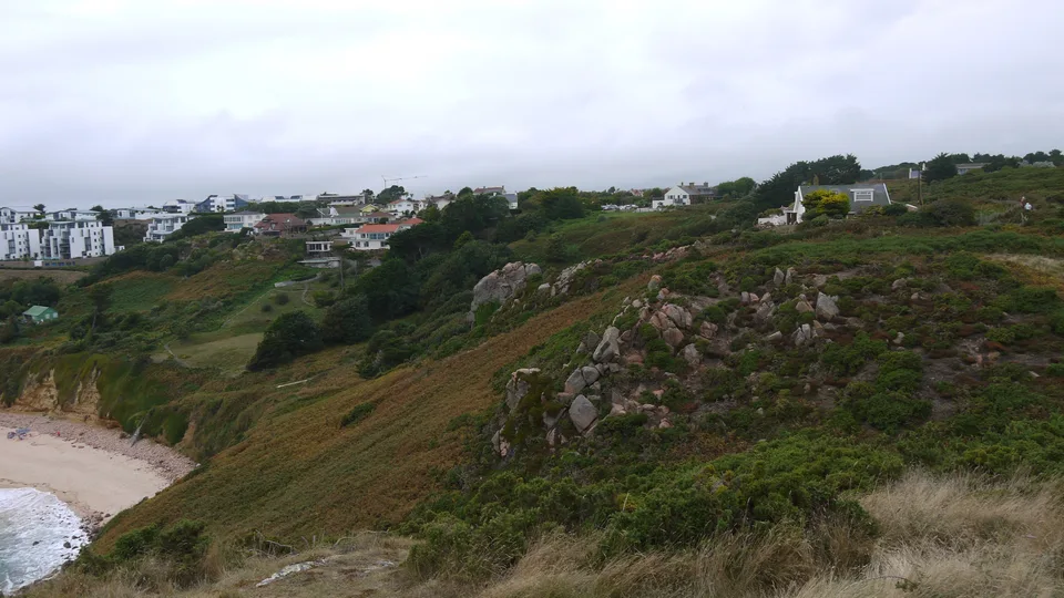 White coastal buildings stand above a steep grassy hillside with trees, rocks and part of a sandy bay below.