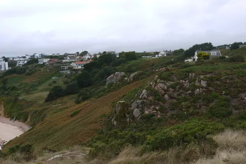 White coastal buildings stand above a steep grassy hillside with trees, rocks and part of a sandy bay below.