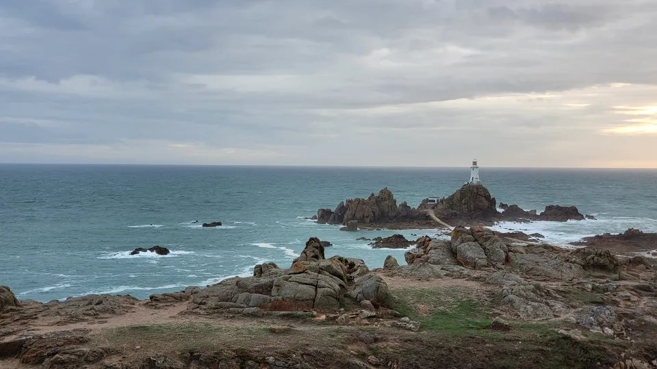 La Corbière's white lighthouse rises from a dark rocky islet, with rough coastal rocks and blue-grey sea in front.