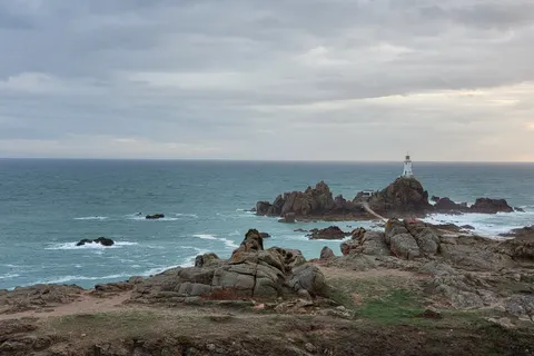 La Corbière's white lighthouse rises from a dark rocky islet, with rough coastal rocks and blue-grey sea in front.