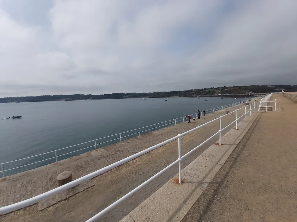 White railings follow a long stone walkway beside calm water, with small figures walking along St Catherine's Breakwater.