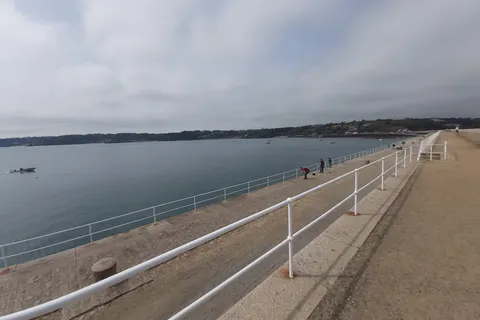 White railings follow a long stone walkway beside calm water, with small figures walking along St Catherine's Breakwater.
