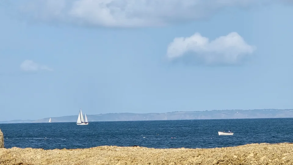A white sailboat and small motorboat cross blue water beyond a narrow strip of pale sand near Grève de Lecq.