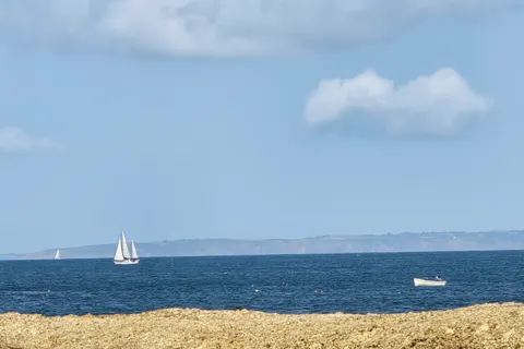 A white sailboat and small motorboat cross blue water beyond a narrow strip of pale sand near Grève de Lecq.