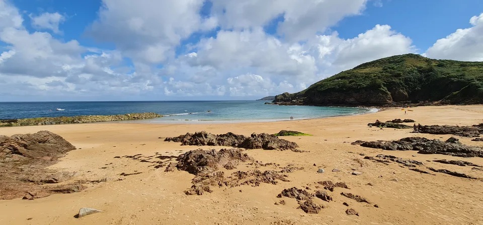 Golden sand and scattered rocks spread across a sheltered cove beneath a wooded headland at Grève de Lecq.