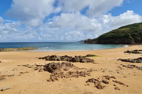 Golden sand and scattered rocks spread across a sheltered cove beneath a wooded headland at Grève de Lecq.