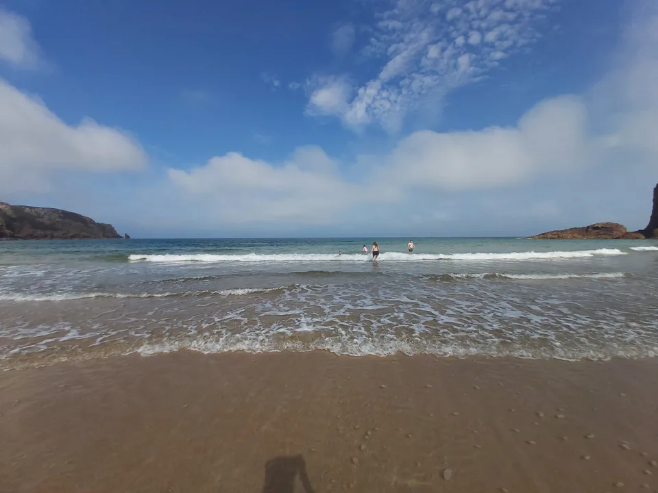 Small waves roll onto a broad sandy shore with a distant person in the water and rocky headlands on either side.