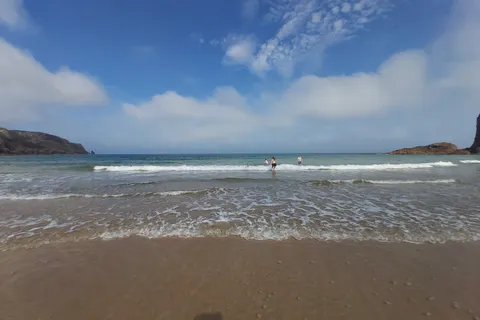 Small waves roll onto a broad sandy shore with a distant person in the water and rocky headlands on either side.