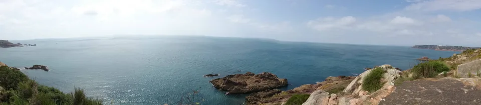 A wide view takes in calm blue sea, rocky outcrops and a grassy cliff edge beneath a hazy sky.