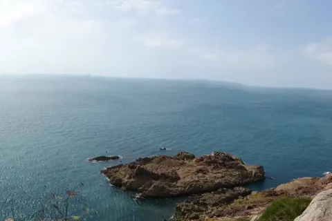 A wide view takes in calm blue sea, rocky outcrops and a grassy cliff edge beneath a hazy sky.