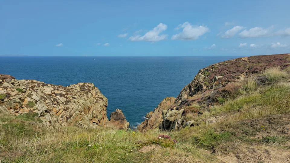 Low rocks and coastal grass border a rocky cleft leading to blue water at Grosnez under a clear sky.
