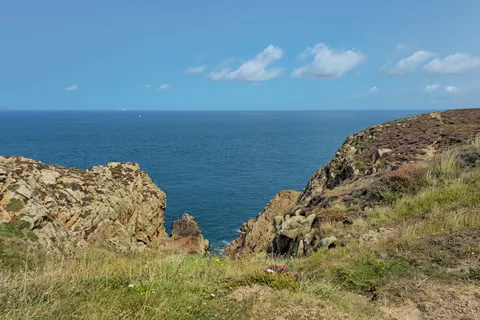 Low rocks and coastal grass border a rocky cleft leading to blue water at Grosnez under a clear sky.